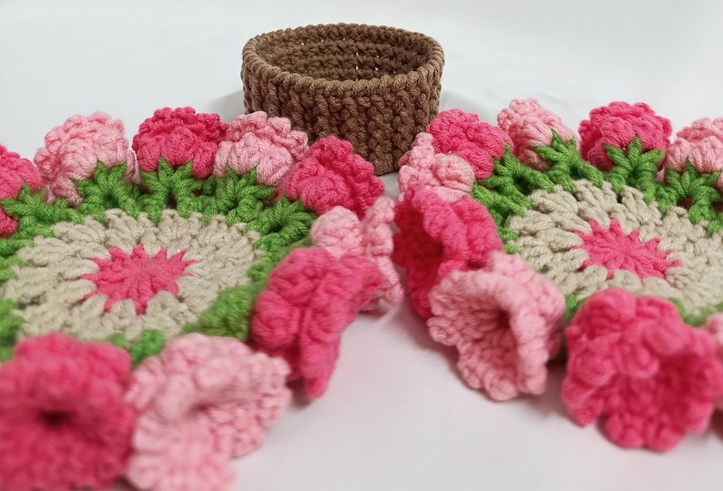 May include: A handmade crochet set featuring a brown basket and floral coasters. The coasters have a pink, green, and cream color scheme, with a flower design. The basket is a solid brown color.