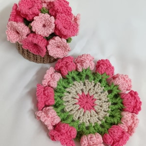 May include: A crocheted floral arrangement in a brown basket and a crocheted coaster. The flowers are in shades of pink and the coaster has a green and beige design. The handmade items are on a white background.