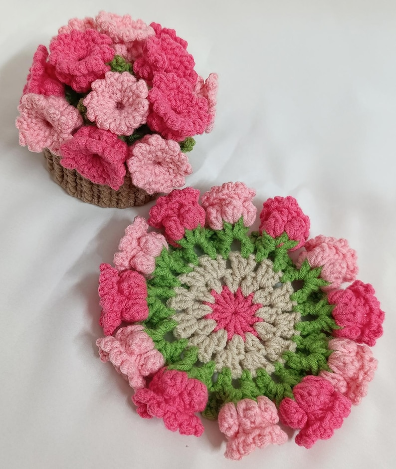 May include: A crocheted floral arrangement in a brown basket and a crocheted coaster. The flowers are in shades of pink and the coaster has a green and beige design. The handmade items are on a white background.