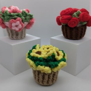 May include: Three crocheted flower arrangements in small baskets. One arrangement features pink and green flowers, another has red roses, and the third has yellow sunflowers with green leaves. The baskets are brown and beige.