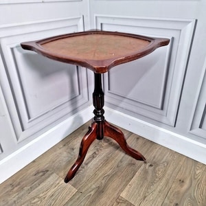 May include: A vintage, dark wood side table with a unique, shield-shaped top. The table has a three-legged base and a central column with decorative detailing. The top is approximately 61 cm wide.