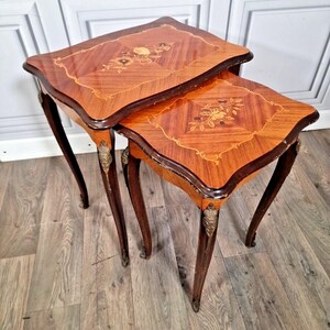 May include: A pair of antique nesting tables in dark wood. Each table has a square top with a floral inlay and curved legs. The edges are trimmed with darker wood. The tables are nested together on a wooden floor. The tables are approximately 50 cm in height.