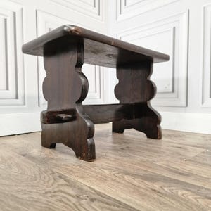 May include: A dark brown wooden stool with a rectangular top and decorative, curved legs. The stool is made of solid wood and has a distressed finish, showing signs of wear. The design is simple and rustic.