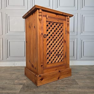 May include: A wooden cabinet with a lattice door and a decorative top. The cabinet is made of light brown wood and has a rectangular shape. The door features a diamond-shaped lattice design. The cabinet is a storage unit.