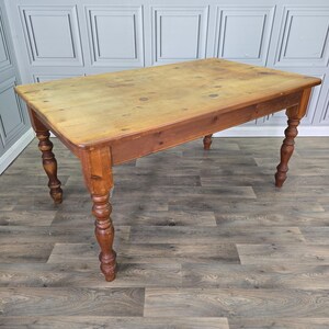 May include: A rectangular wooden table with a natural light brown finish on the top surface and a darker brown tone on the frame and legs. The table features four turned legs with a rounded, decorative profile and a smooth, flat tabletop surface.