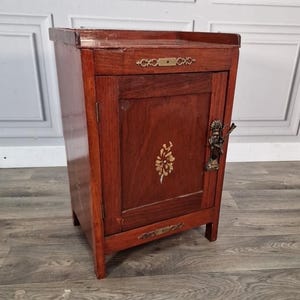 May include: A small, antique wooden cabinet with a single door. The cabinet is a rich, reddish-brown colour with brass hardware and a decorative gold floral design on the door. The cabinet is approximately 61 cm tall.