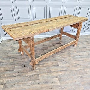 May include: A distressed wooden table with a rectangular top. The table is light brown and has a two-plank top. The legs are angled and sturdy. The table's dimensions are suitable for a variety of uses. The table's length is approximately 180 cm.