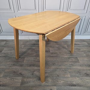 May include: A light-coloured wooden drop-leaf table with four straight legs. The table has an oval shape with one leaf extended. The table is approximately 100 cm long and 75 cm wide when fully extended.