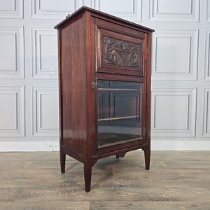 May include: A dark brown wooden cabinet with a glass door and carved floral details. The cabinet has a rectangular shape, stands on four legs, and features a decorative carved panel above the glass door. The cabinet is approximately 152 cm tall.