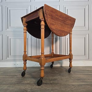 May include: A vintage wooden tea trolley with a drop-leaf design. The trolley is made of light brown wood and features turned legs, a lower shelf, and small black wheels. The top is partially open, revealing the interior.