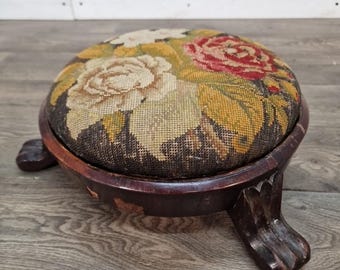 May include: A round footstool with a dark brown wooden frame and three carved legs. The top features a needlepoint fabric cushion displaying a floral pattern with large white and red roses against a dark brown background with green leaves.