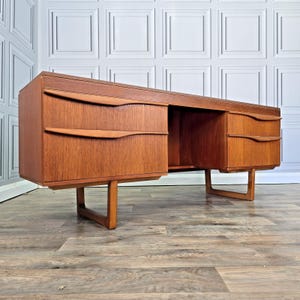May include: A teak wood desk featuring a rectangular top, a left-side pedestal with two drawers, and a right-side pedestal with two drawers. Each drawer is equipped with a sculpted, integrated wooden handle. The desk is supported by four legs.