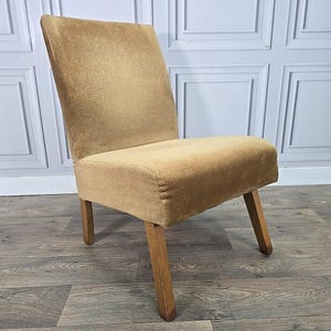 May include: A vintage chair upholstered in tan velvet. The chair features a simple design with a rectangular back and seat, supported by four wooden legs. A mid-century modern style chair, suitable for a living room or bedroom.