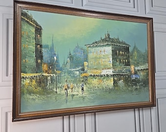May include: An oil painting of a European city scene, framed in a dark wood. The artwork depicts buildings, streetlights, and figures in muted greens, blues, and yellows, creating a vintage aesthetic. The painting is mounted on a white wall.