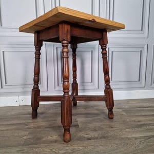 May include: A small, antique wooden table with a square top and turned legs. The tabletop is a light, natural wood colour, while the legs and frame are a darker, stained wood. The table is approximately 61 cm tall.