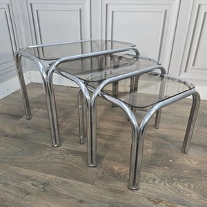 May include: A set of three nesting tables featuring polished chrome tubular metal frames and rectangular smoked glass tops. The tables are arranged in a cascading formation, showcasing the reflective metallic finish and the translucent dark glass.