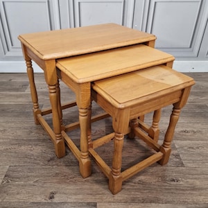 May include: A set of three light brown wooden nesting tables with rectangular tops and smooth surfaces. Each table features four turned legs connected by horizontal stretchers near the base, allowing the smaller tables to slide under the largest one.