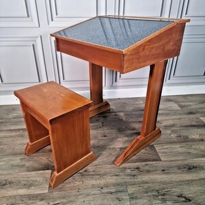 May include: A vintage wooden desk and stool set. The desk has a slanted top with a textured, dark grey surface. Both the desk and stool are made of stained wood, displaying a warm, reddish-brown colour. The desk features a distinctive angled leg design.
