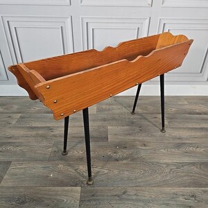 May include: A vintage wooden planter box with a scalloped edge, supported by four black metal legs. The planter is made of light brown wood and is secured with small metal rivets. A decorative piece for plants.