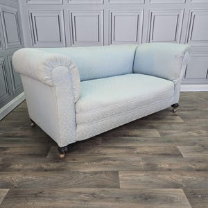 May include: A light blue sofa featuring a subtle damask pattern, rounded scroll arms, and a single bench seat cushion. The piece is supported by dark wooden legs with small metal castors at the base, providing a low-profile seating option.