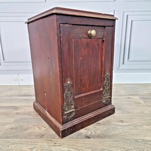 May include: A dark brown, antique wooden cabinet with a single door and a small, round, cream-coloured knob. The cabinet has decorative metal hinges and a slightly distressed finish, suggesting age and use. The cabinet is rectangular and sits on a wooden floor.