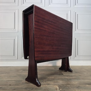 May include: A dark mahogany wood drop-leaf table shown in its folded, upright position. The table features a smooth, rectangular surface with a visible wood grain pattern and is supported by two sturdy, curved legs on each side of the base.
