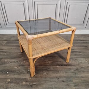 May include: A square side table constructed from light brown natural rattan with a dark smoked glass top. The table features a lower shelf made of woven rattan cane material and four sturdy legs reinforced with wrapped rattan bindings at the joints.