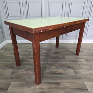 May include: A rectangular table featuring a light green laminate top surface and a dark brown wooden frame with four straight legs. The wooden edges show signs of wear and finish loss, revealing the natural grain and texture of the wood construction.