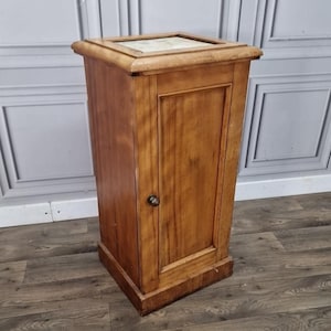 May include: A tall, wooden cabinet with a single door and a marble top. The cabinet is made of light brown wood and has a simple, classic design. The door has a small, round knob. The cabinet is approximately 76 cm tall.