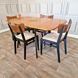 May include: A vintage dining set comprising a light brown oval table and four chairs. The chairs have black frames, cream-colored seat cushions, and curved wooden backs. The table has a similar light brown finish, typical of mid-century design.