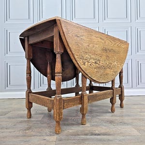 May include: A vintage, wooden drop-leaf table with a light brown finish. The table has a round top with a drop-leaf design, supported by turned legs and a rectangular base. The table's dimensions are approximately 76 cm in height and 91 cm in diameter when fully extended.
