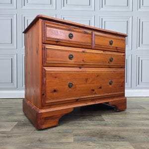 May include: A wooden chest of drawers in a warm, reddish-brown hue. It features four drawers with dark, round knobs and a slightly curved base. The wood grain is visible, giving it a rustic appearance. The chest is approximately 100 cm wide.