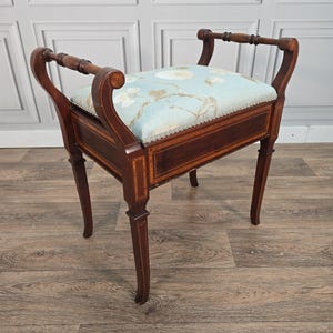 May include: A wooden bench with a rectangular seat upholstered in light blue fabric featuring a white floral pattern. The frame is dark brown wood with inlaid light wood detailing, curved armrests, and four straight legs with tapered feet.