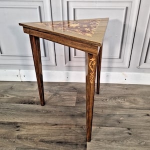 May include: A triangular side table with a dark wood finish and decorative inlay. The tabletop features a floral design in lighter wood tones. The table has three slender legs, each with a decorative accent. The table is approximately 61 cm tall.