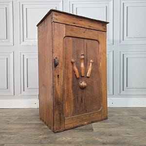 May include: A vintage wooden cabinet with a single door and a dark finish. The door has three decorative wooden features and a round knob. The cabinet has a metal handle and a keyhole, suggesting it is lockable. The wood grain is visible throughout.