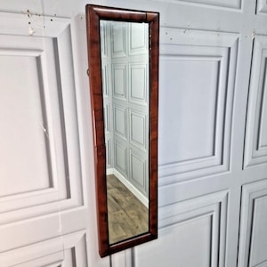 May include: A long, rectangular mirror with a dark brown wooden frame. The mirror is mounted on a white wall. The frame has a wood-grain appearance. The mirror reflects a room with white paneling and a wooden floor.