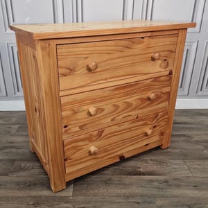 May include: A light brown wooden chest of drawers featuring three horizontal drawers, each equipped with two round wooden knobs. The piece displays a natural wood grain pattern throughout its rectangular frame and stands on four short, sturdy legs.