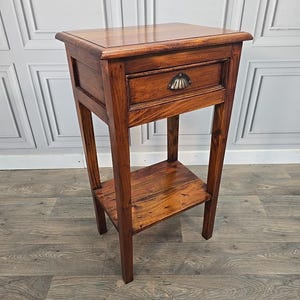 May include: A small, wooden side table with a drawer and a lower shelf. The table is a rich, dark brown colour with a brass-coloured drawer pull. The table is approximately 71 cm tall, 46 cm wide, and 36 cm deep.