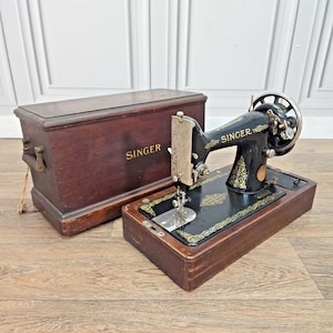 May include: A black Singer sewing machine featuring gold filigree patterns and a silver needle plate, mounted on a rectangular wooden base. Beside it sits a matching dark brown wooden carrying case with the word SINGER printed in gold lettering.