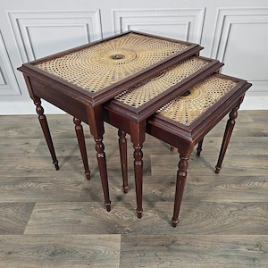 May include: A set of three nesting tables. Each table has a dark wood frame and a woven cane top. The rectangular tops have a circular woven design. The tables have slender, turned legs.