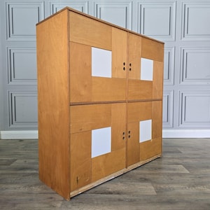 May include: A tall wooden cabinet featuring four doors arranged in a grid. Each door is constructed from light brown wood panels with a central square white inlay. The cabinet has a rectangular frame and small circular cutouts serving as door handles.