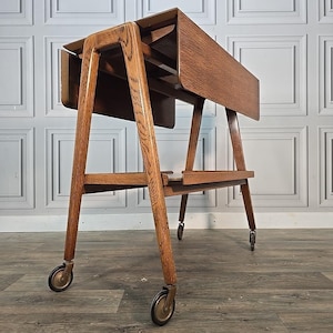 May include: A vintage wooden tea trolley with a drop-leaf design. The trolley has a warm, medium-brown finish, supported by angled legs and small caster wheels. The tabletop is rectangular when open and folds down for storage.
