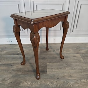 May include: A small, antique wooden side table with a glass top. The table has four curved legs with carved shell details. The wood is a warm brown colour, and the table is likely used for decorative purposes.