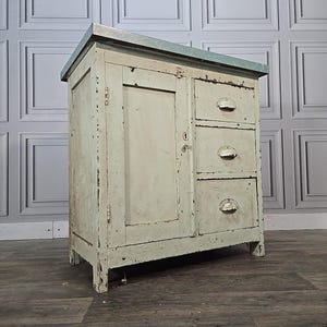 May include: A distressed, off-white cabinet with a metal top. The cabinet has a single door and three drawers with rounded pulls. The paint is chipped, giving it a vintage look. It stands on a wooden floor.