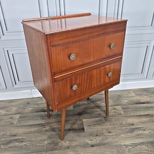 May include: A two-drawer wooden bedside cabinet in a reddish-brown hue. The cabinet has decorative brass knobs and a thin gold accent. It stands on four slender, tapered legs, reflecting a classic mid-century modern design. Dimensions are approximately 60cm high.