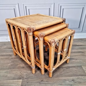May include: A trio of nesting tables crafted from light brown rattan. Each table features a square woven top and curved, arched legs. The tables are designed to fit inside each other, providing a space-saving furniture solution.