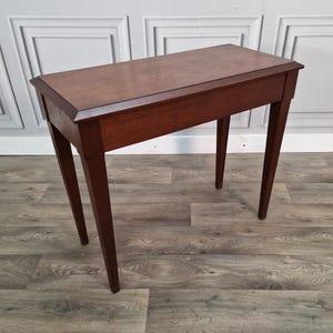 May include: A rectangular wooden console table with a smooth, dark brown finish. The table features a flat top surface with bevelled edges and four straight, tapered legs that extend downwards from the corners of the rectangular frame.