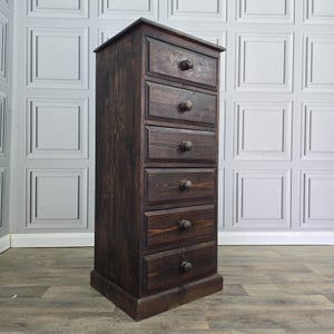 May include: A tall, narrow chest of drawers crafted from dark stained wood with a visible grain. The unit features six stacked drawers, each equipped with a single round wooden knob centred on the front panel, standing on a solid base.
