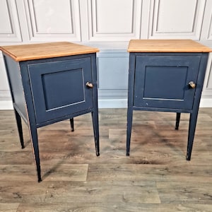 May include: A pair of matching bedside tables featuring a dark navy blue painted finish on the frame and a natural light brown wood top. Each table has a single cabinet door with a small round knob and stands on four straight tapered legs.