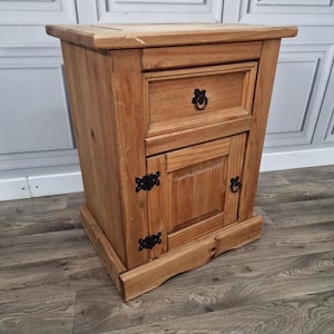 May include: A wooden bedside table with a drawer and a small cupboard door. The table is made of light-coloured wood and has black metal hardware. The drawer has a round handle, and the cupboard door has a matching handle. The table is a rustic style.